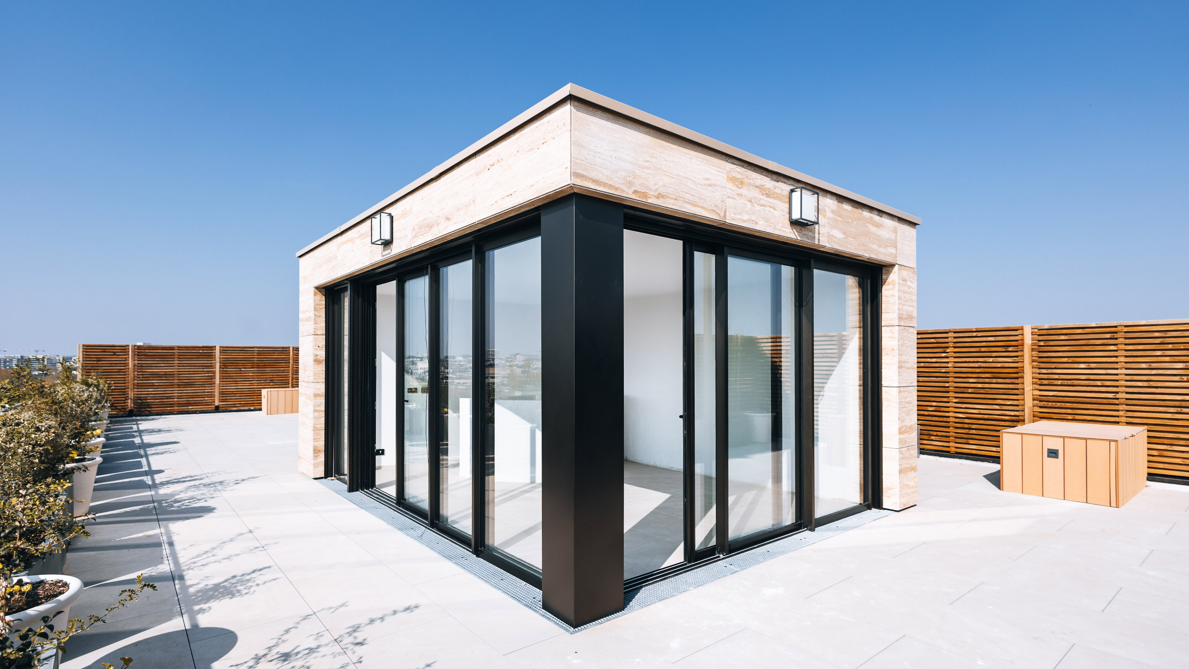 Extension moderne sur toit-terrasse parisien avec larges baies coulissantes et habillage en pierre naturelle sous un ciel bleu d&eacute;gag&eacute;. par maxime decarsin photographe
