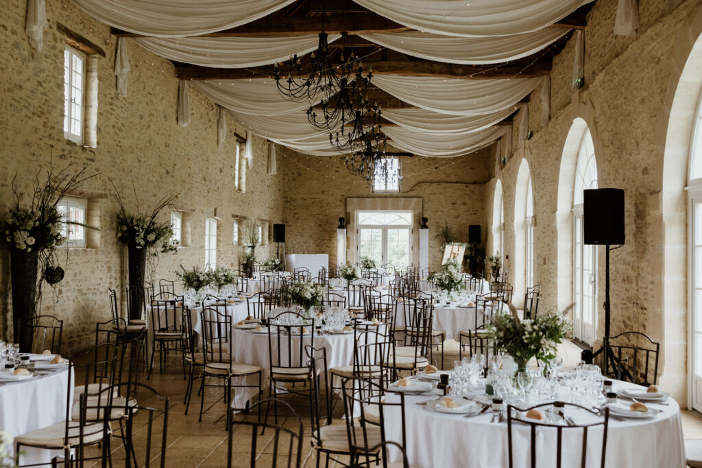 Salle de réception aux murs en pierre, tables rondes dressées et tissu blanc drapé au plafond